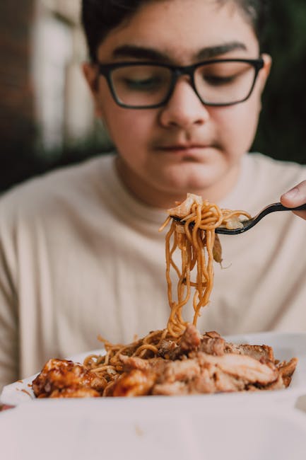 A teenage boy with glasses enjoys a takeout meal of noodles and chicken, captured in a close-up shot.